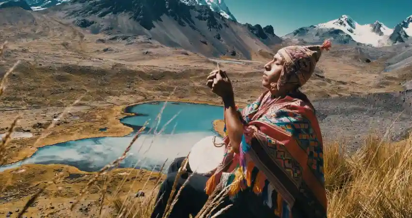 A woman in traditional clothing sits on a hill, gazing at a serene lake during a spiritual journey in Peru.