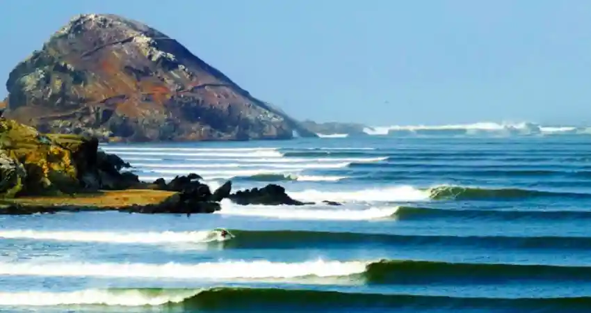 A large rock stands in the ocean at Chicama, Peru, with waves crashing around it, creating a dynamic coastal scene.
