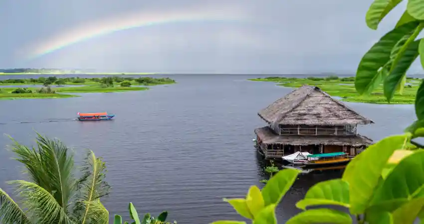 A vibrant rainbow arcs over a small house by the water, showcasing the beauty of a luxury Amazon tour in Peru.