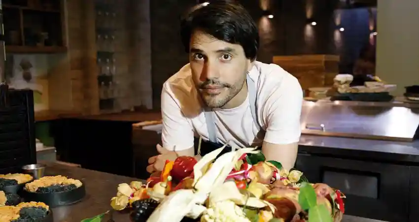 A man stands before a large plate of food at a chef's table in Cusco, showcasing a vibrant culinary presentation.