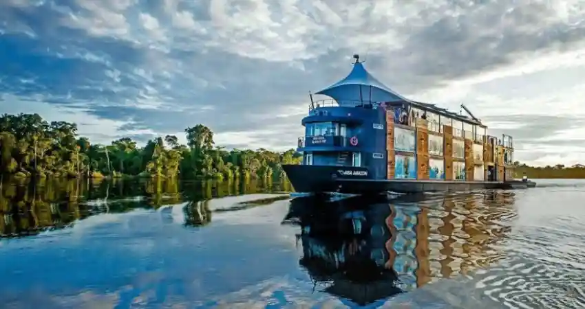A luxury houseboat floats on calm water, surrounded by lush trees, promoting serene tours in Peru.