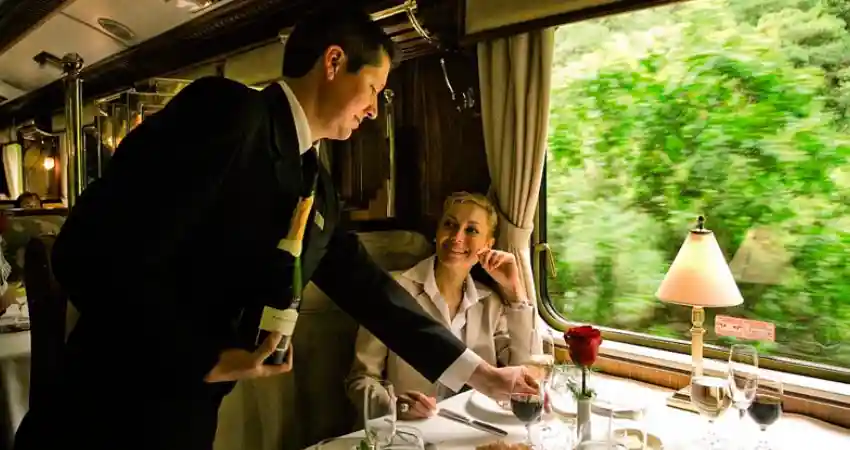 A man in a suit serves wine to a woman on a luxury train, highlighting an elegant experience on a Machu Picchu tour.