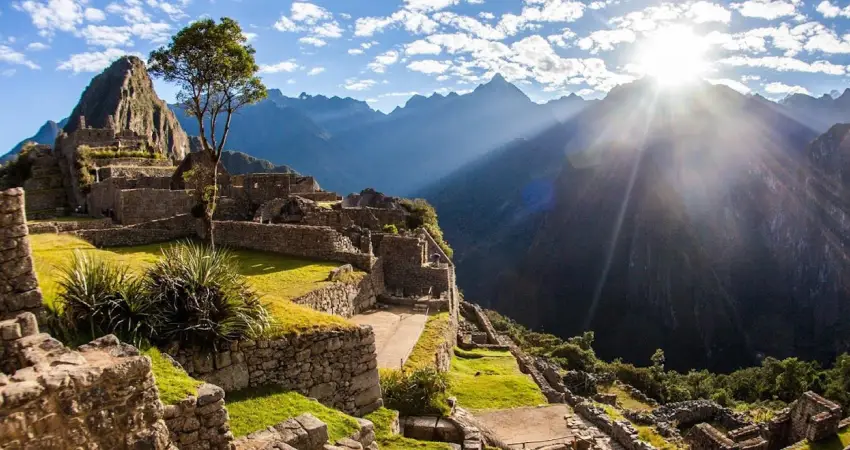 The sun rises brightly over Machu Picchu, illuminating the ancient ruins and surrounding mountains.