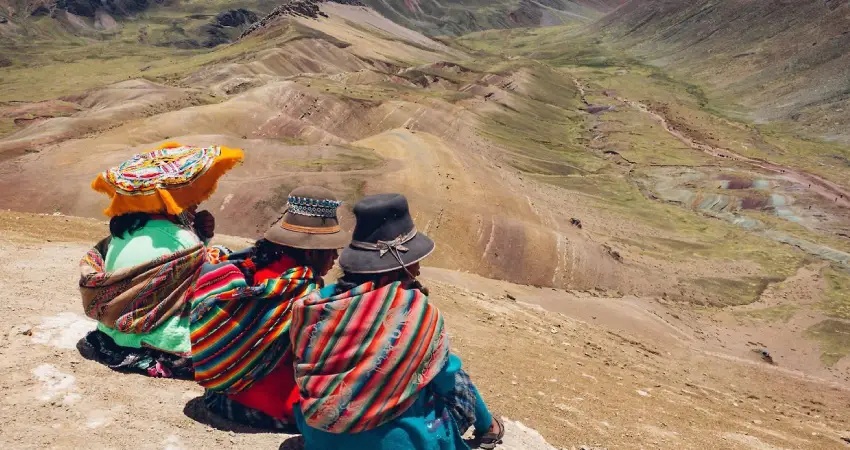 Three women in colorful clothing sitting on a mountain during a custom trip to Perú, enjoying the scenic view.