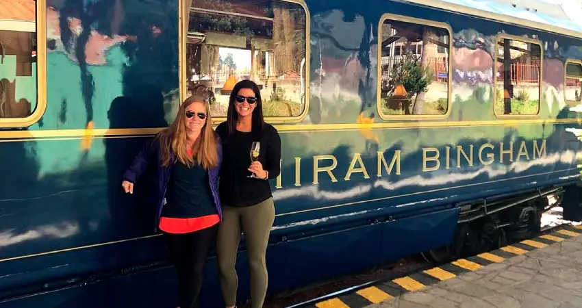 Two women pose for a photo in front of the Lima to Cusco Luxury Train, smiling against a scenic backdrop.