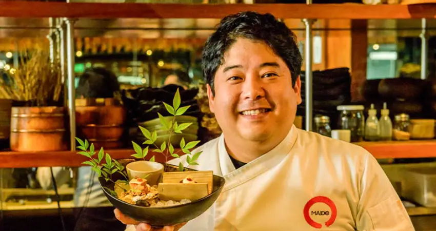 Chef at Maido Restaurant in Lima holds a bowl of food, with a shelf of ingredients visible in the background.