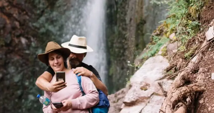 A man and woman take a selfie in front of Poc Poc Waterfall in Sacred Valley, surrounded by lush greenery.