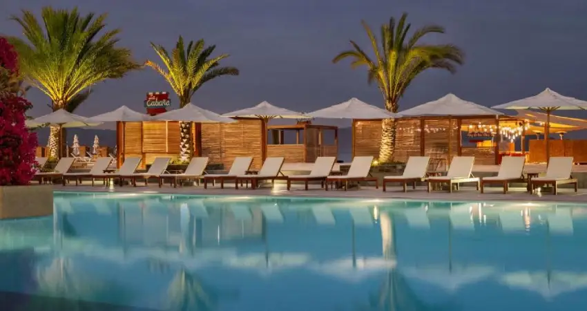The illuminated pool at The Legend Paracas Resort beach club, surrounded by palm trees, glows under the night sky.