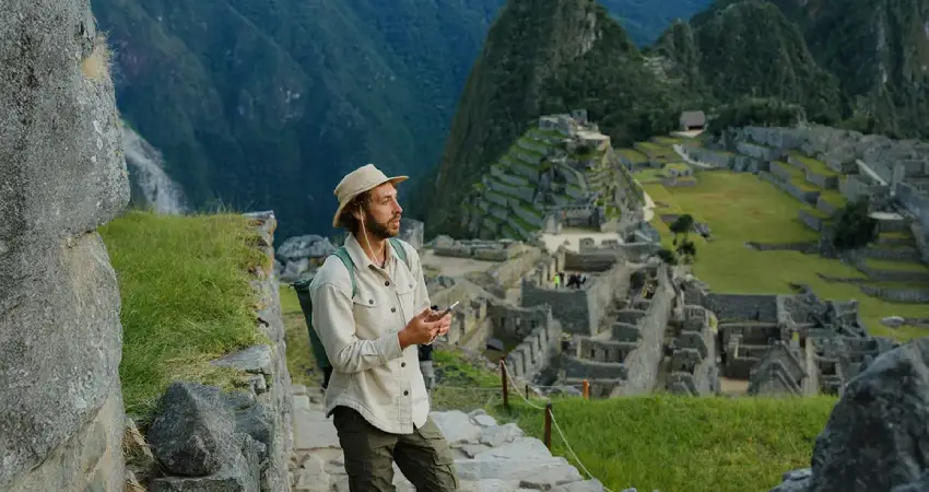 A man in a hat stands on a stone wall, gazing at the ancient ruins of Machu Picchu in Peru.
